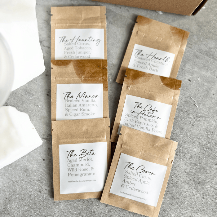 Thrifted Shelf Creepy vs. Cozy Scent Sampler Pack featuring six labeled kraft pouches — The Haunting, The Manor, The Bite, The Hearth, The Café in Autumn, and The Coven — laid out on a gray surface.