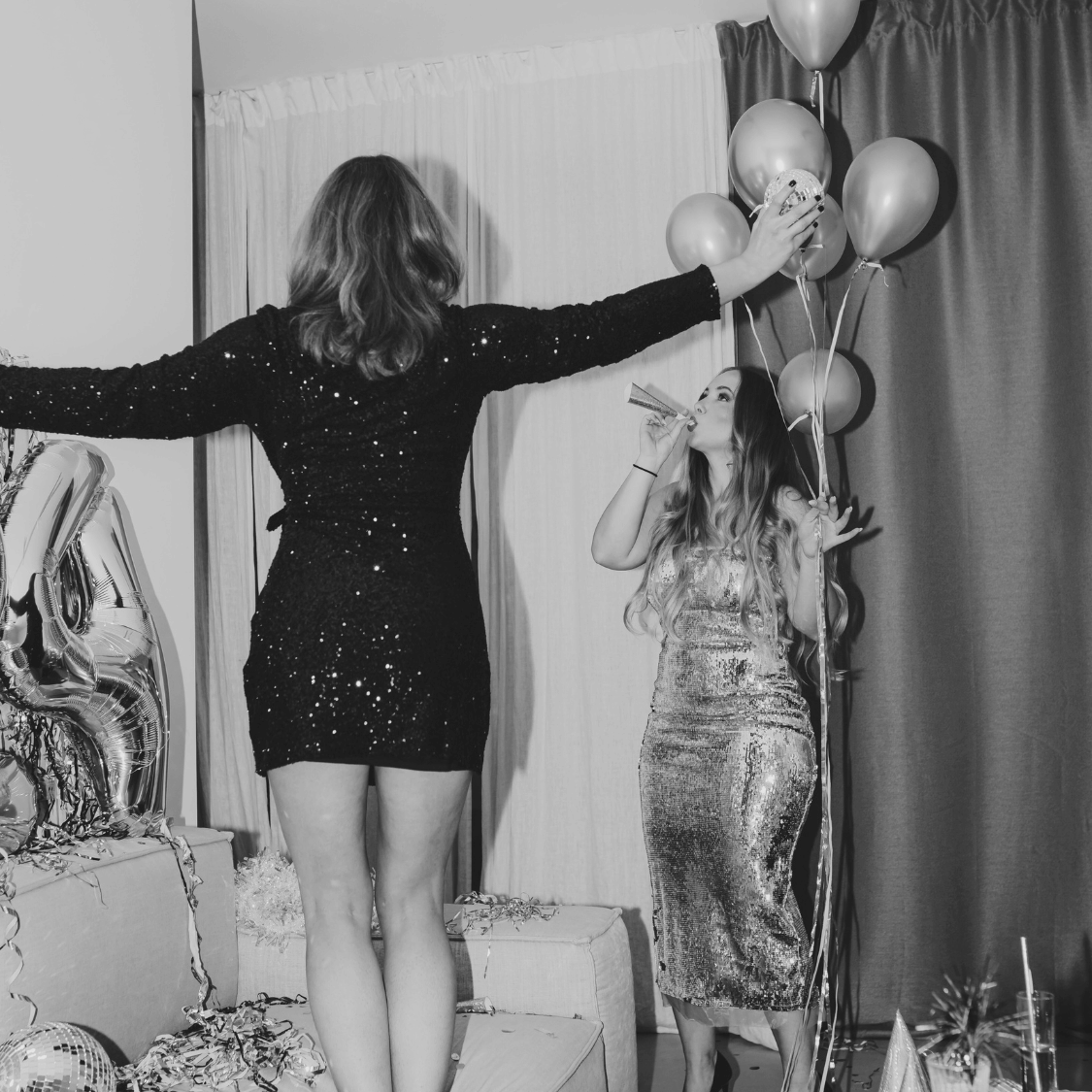 Two women in sparkly dresses standing in a room with balloons and decorative items.