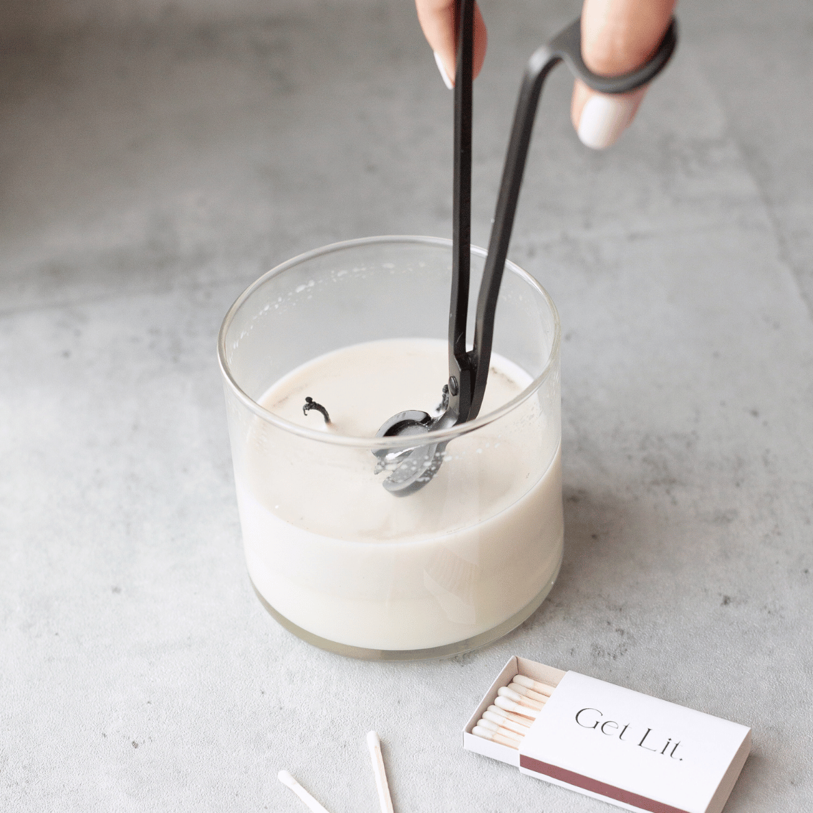 Candle being lit with tongs on a gray surface, with a box of matches labeled 'Get Lit' nearby.