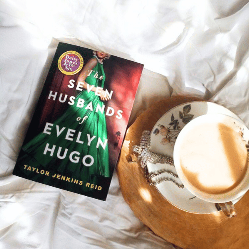 The Seven Husbands of Evelyn Hugo by Taylor Jenkins Reid on white bedding with a cup of coffee and pearls, bestselling historical fiction novel about love, fame, and secrets.