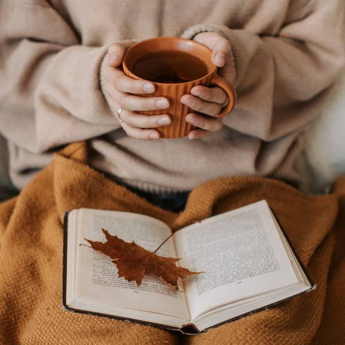 Person holding a warm mug of tea while reading a book, with a fall leaf resting on the pages, wrapped in a cozy autumn blanket.