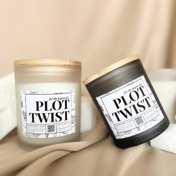 Two matte black candles labeled “Plot Twist” with wooden lids, one standing upright and one resting on its side, styled on beige fabric with chunks of white wax.