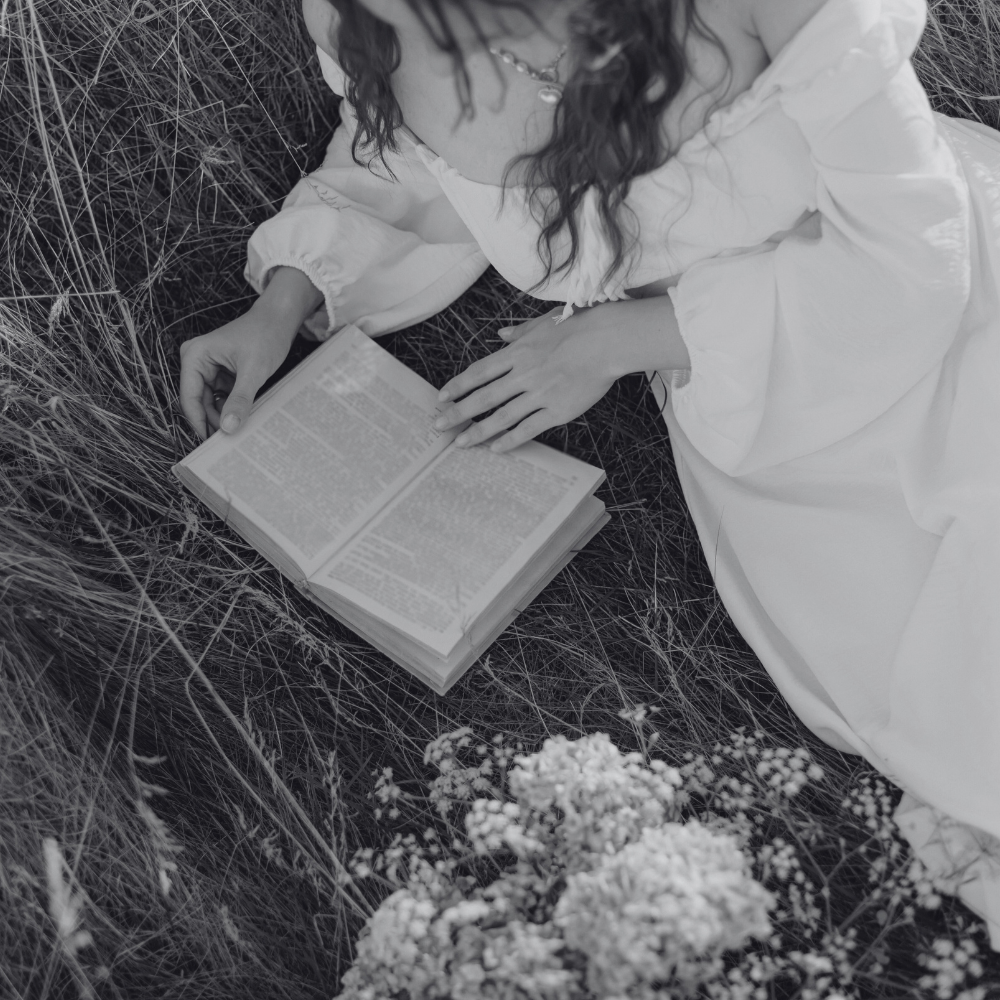 Woman in a flowing white Regency-style dress reading a vintage book while sitting in tall grass, romantic aesthetic for Regency romance readers.