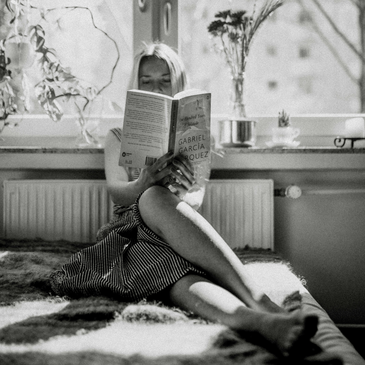 Woman sitting on a bed in natural sunlight reading “One Hundred Years of Solitude” by Gabriel García Márquez, representing classic and contemporary fiction.