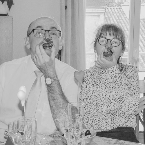 Playful couple wearing glasses and holding cherries by their mouths in a humorous, romantic gesture during a cozy meal, symbolizing fun and love for romance book fans.