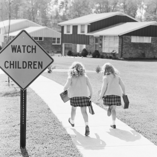 Two young girls in matching plaid skirts and cardigans walking to school with lunchboxes, passing a “Watch Children” sign in a suburban neighborhood.